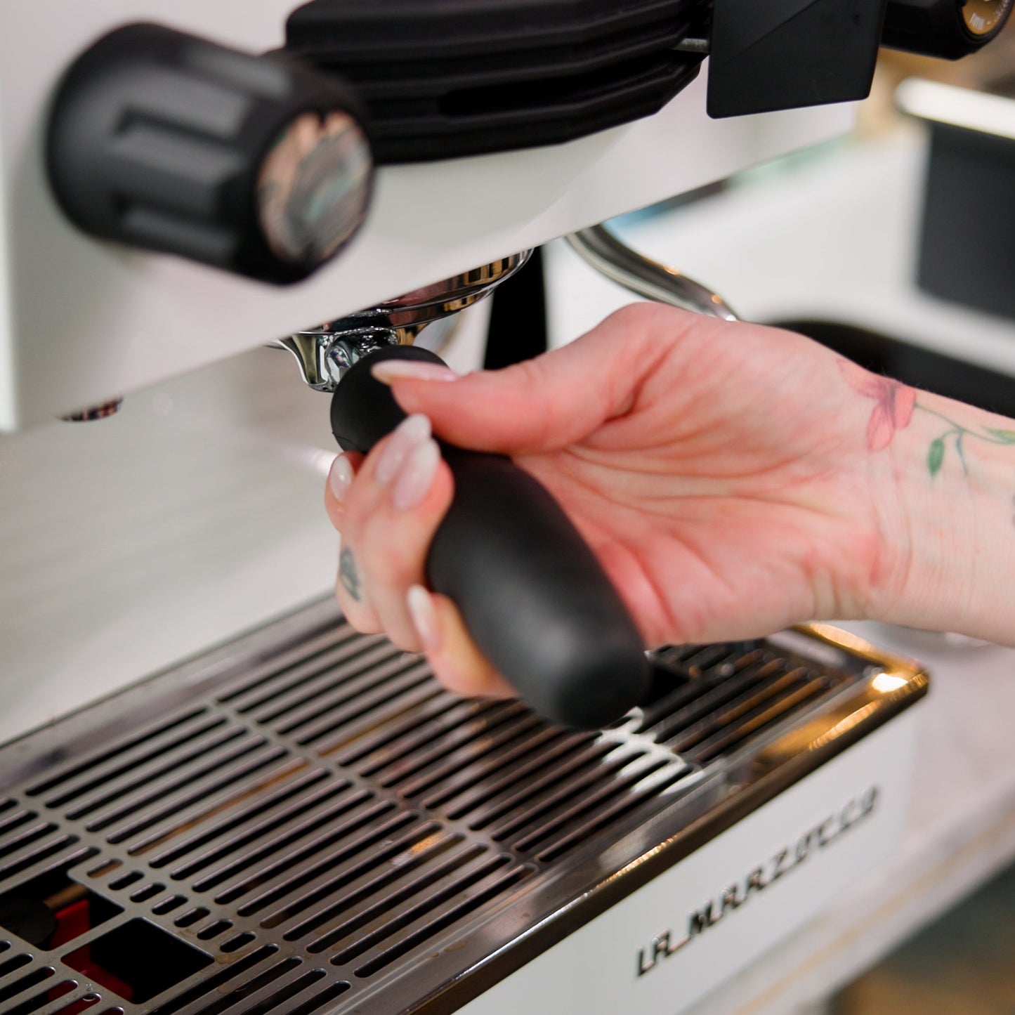 A close-up of a tattooed hand holding the portafilter handle under the espresso spout of a white and stainless steel espresso machine.